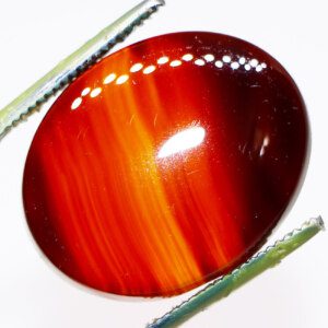 Close-up photo of a Red Line Agate loose gemstone held in tweezers, highlighting its red stripes and unique patterns.